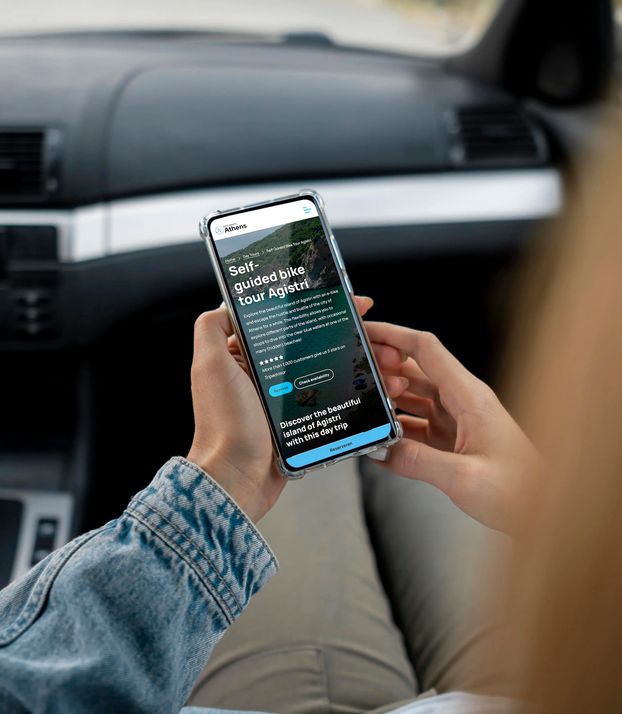 Iemand in de bijrijderstoel van een auto houdt een smartphone vast die de website van Meet in Athens bekijkt.